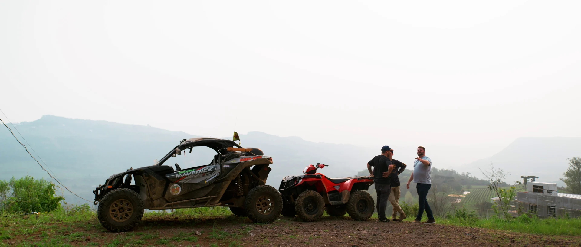Veículos off-road enlameados estacionados no alto de um morro enquanto três homens conversam, com a serra enevoada ao fundo.