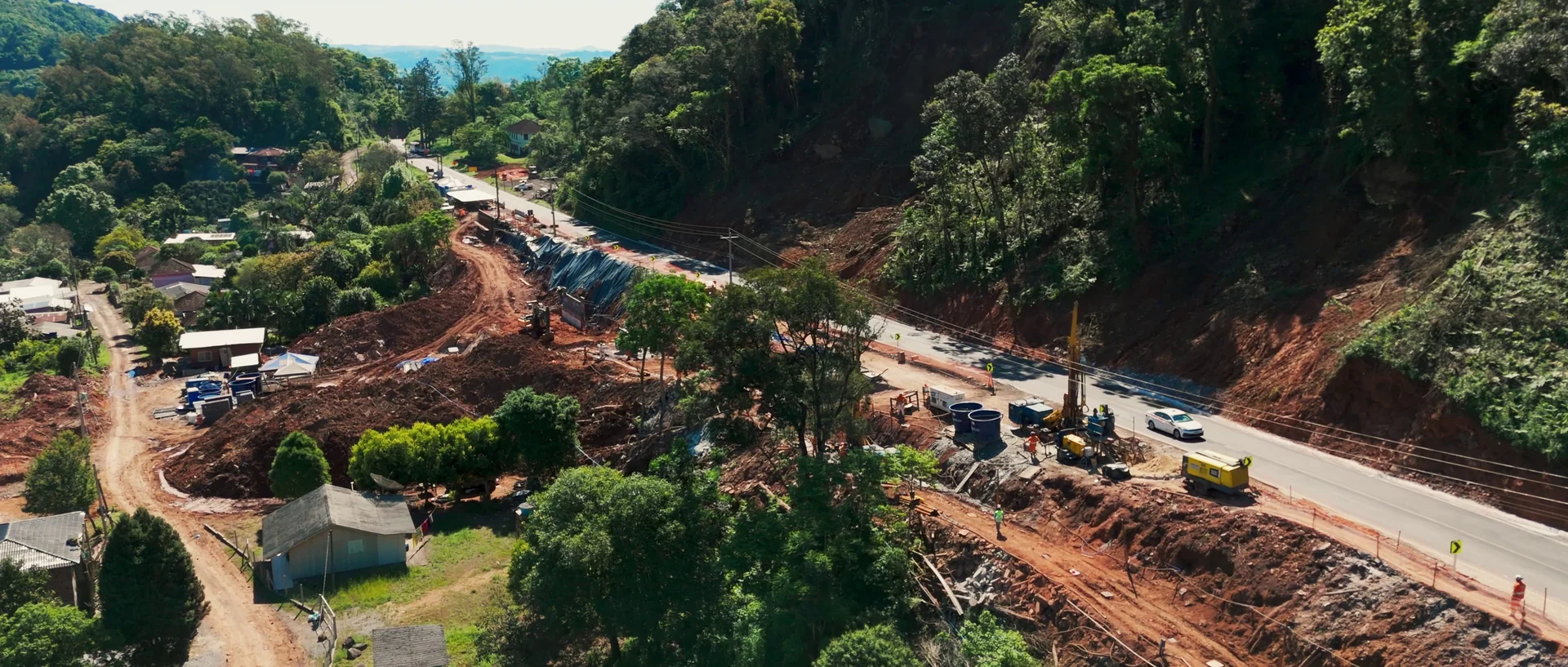 Vista aérea de uma rodovia em obras ao pé de uma encosta de terra, com máquinas e caminhões trabalhando na reconstrução.