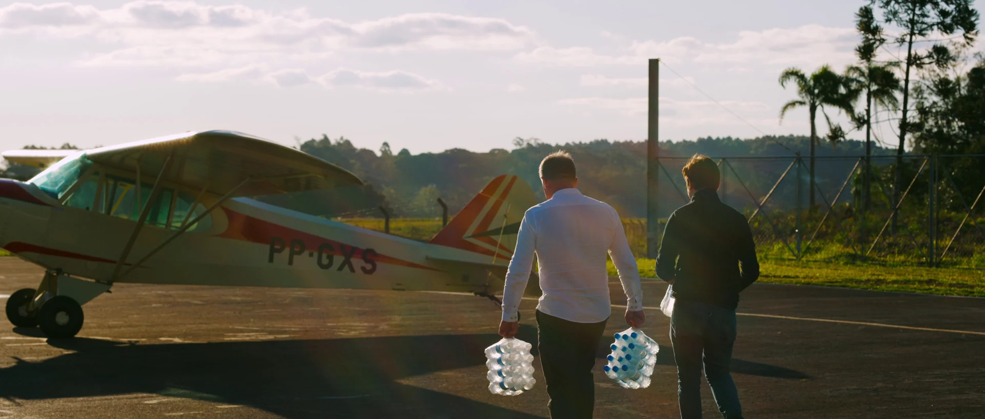 Dois homens de costas carregam fardos de garrafas de água em direção a um pequeno avião na pista do aeroclube, sob a luz do fim de tarde.