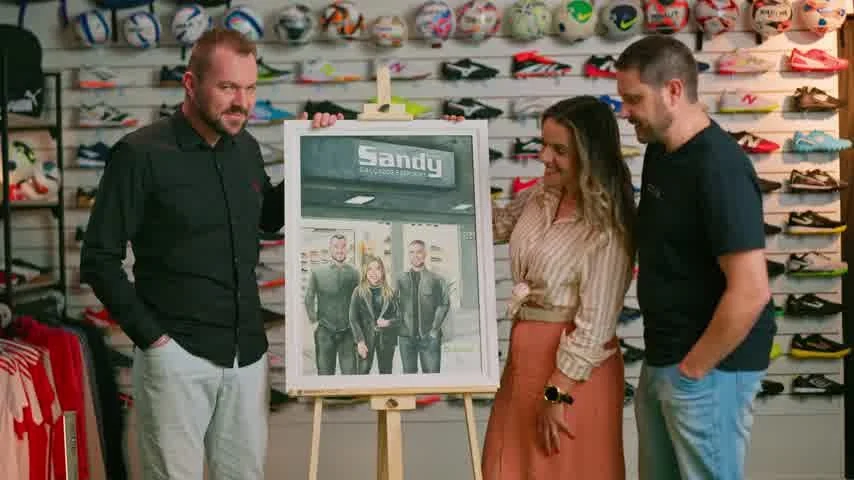 Dois homens e uma mulher posam ao lado de um quadro com a fachada da Sandy Calçados, diante de uma parede de chuteiras e bolas.
