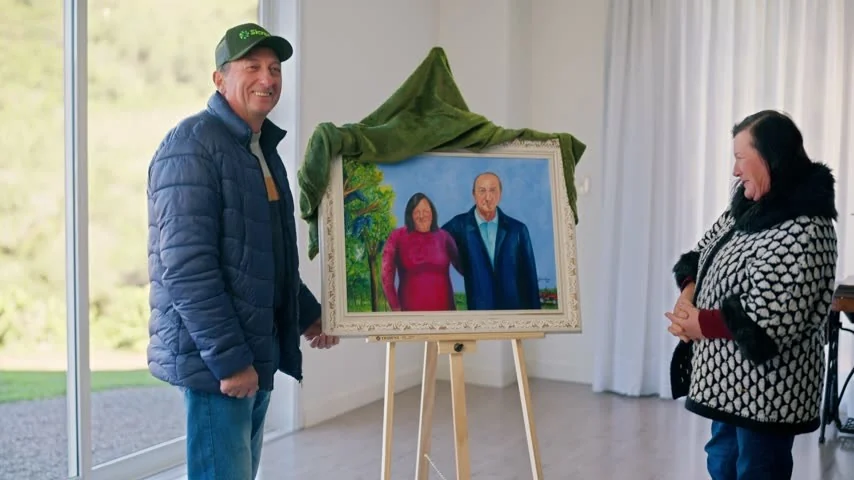 Nivaldo Matana, de boné e jaqueta azul, segura o quadro recém-revelado com o retrato do casal enquanto Vera Matana sorri ao lado, numa sala clara.