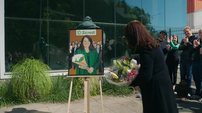 Rejane Fontana, com um buquê de flores, olha para o próprio retrato num cavalete enquanto colegas aplaudem na entrada da sede da cooperativa.