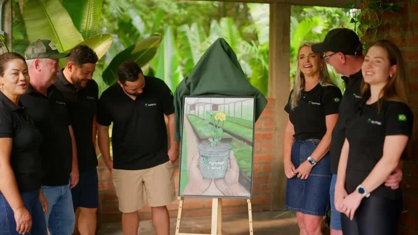 A família da Agriplugs, de camisetas pretas, se reúne sob um telhado de madeira em volta de um quadro ainda coberto por um pano verde, entre folhagens.