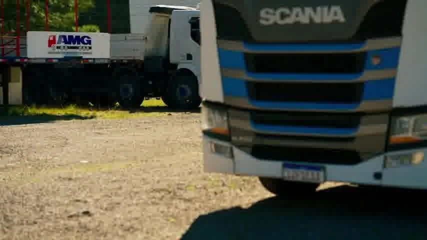 Frente de um caminhão Scania branco em primeiro plano, com outro caminhão e sua carreta estacionados no pátio de terra ao fundo.