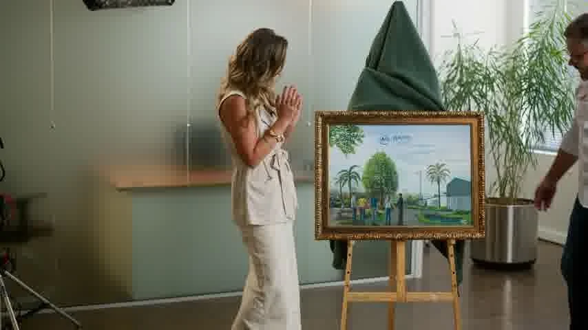 Ana Thaís, embaixadora do Juntos pelo Caí, de roupa clara, junta as mãos emocionada diante do quadro do projeto, apoiado num cavalete.