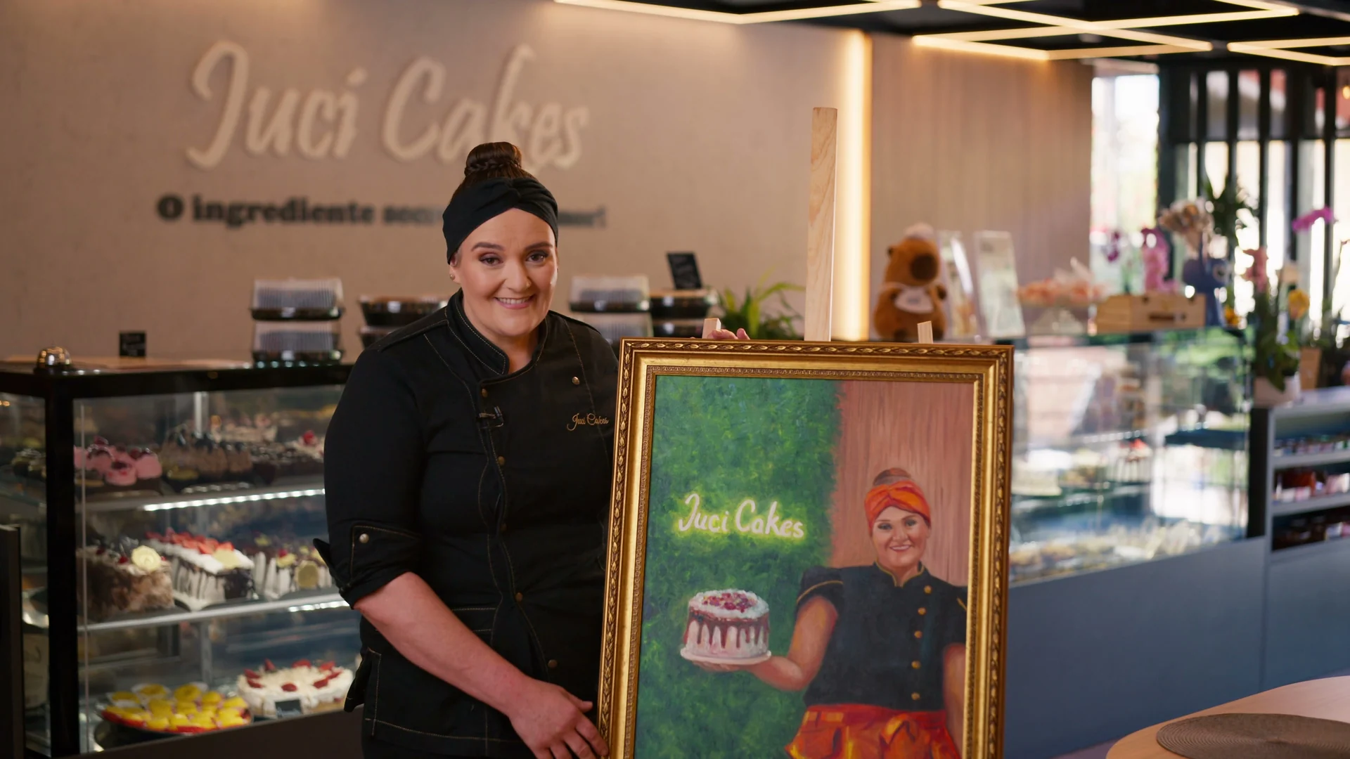 Juciana Fabrin Vieira, de dólmã preto e faixa no cabelo, sorri ao lado do quadro que a retrata segurando um bolo, diante das vitrines de doces da Juci Cakes.