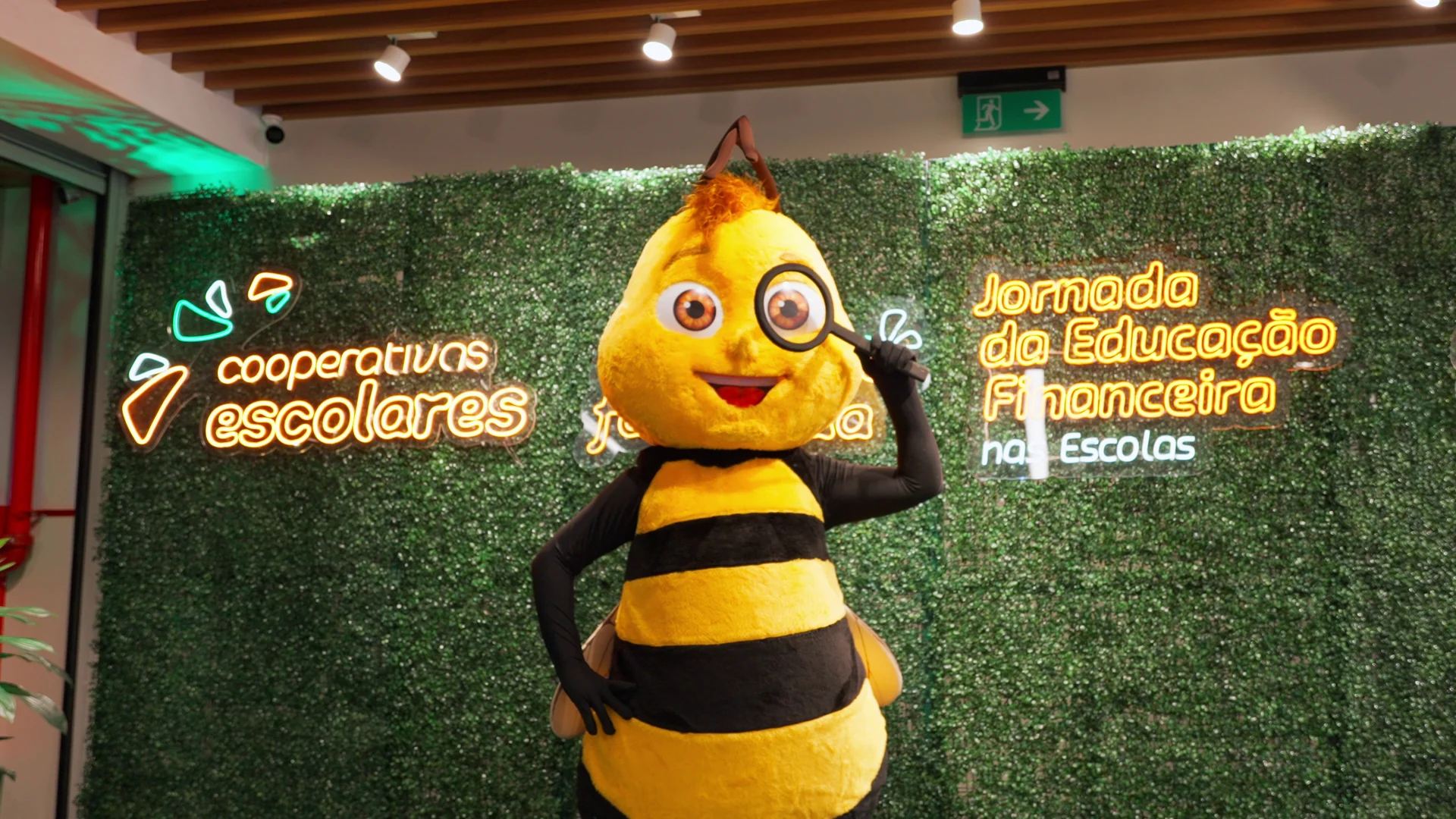 Mascote de abelha com lupa posa diante de uma parede verde com letreiros em neon de Cooperativas Escolares e Jornada da Educação Financeira nas Escolas.