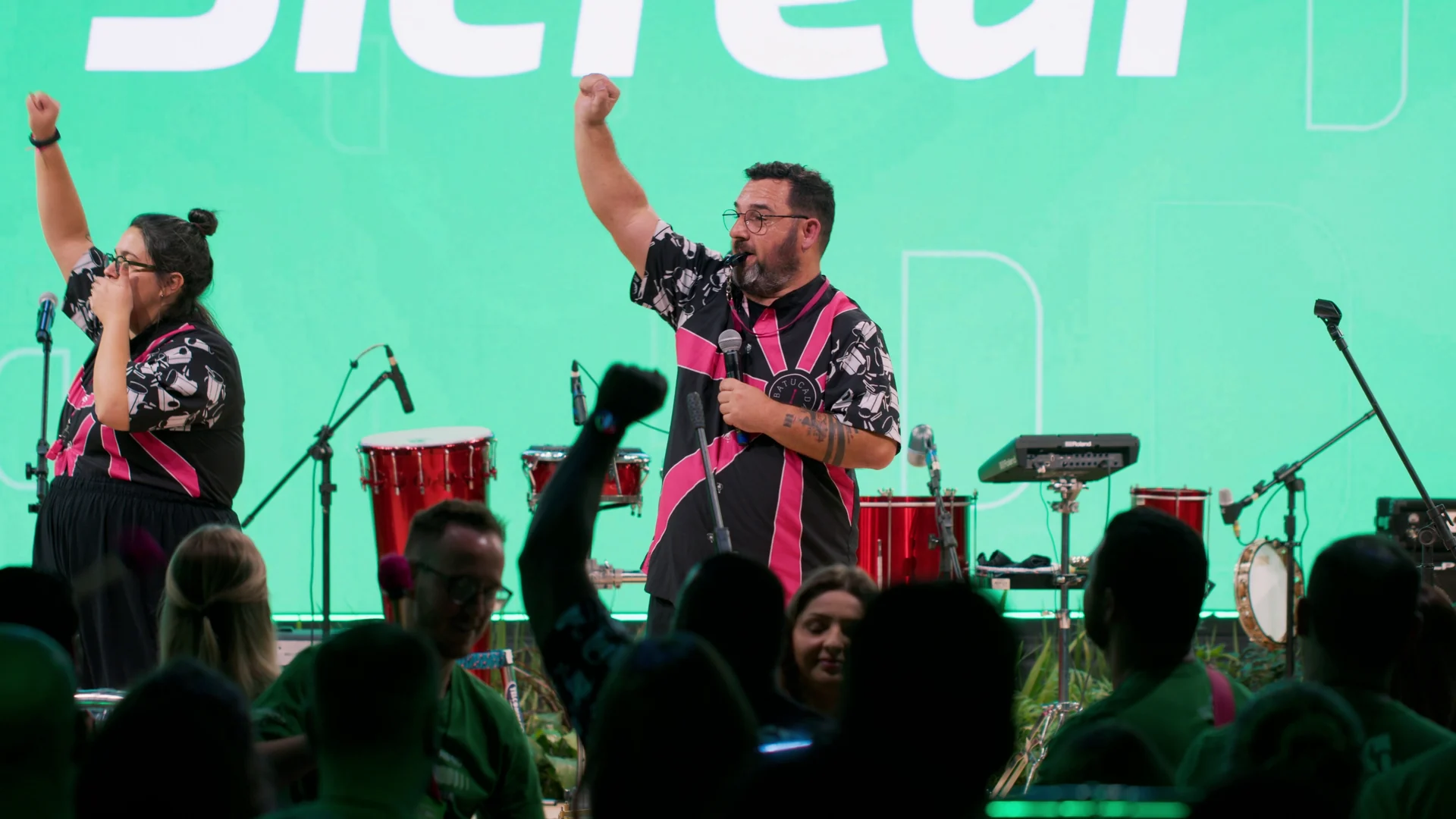 Dois percussionistas de camisa rosa e preta erguem o punho no palco verde, com tambores vermelhos atrás e o público em silhueta à frente.