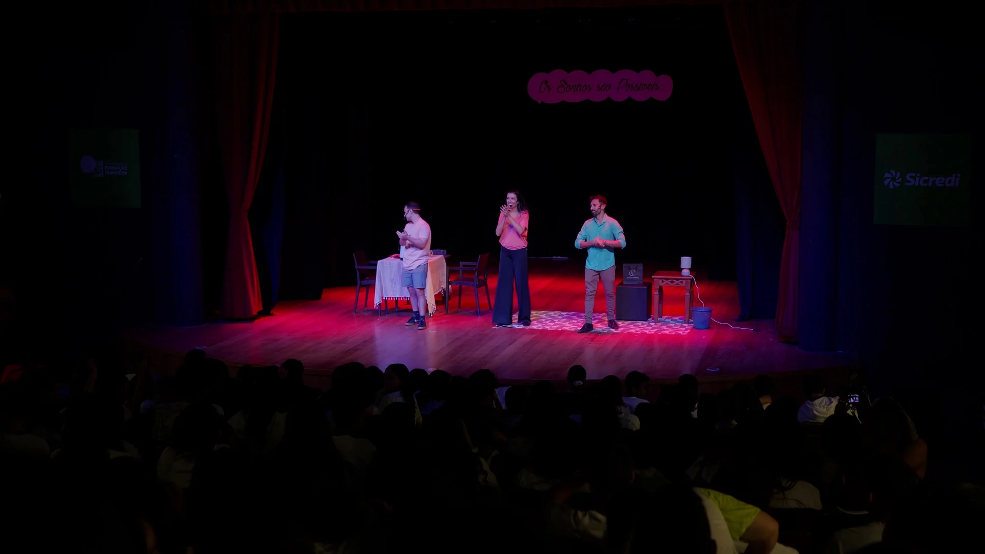 Três atores se apresentam num palco de teatro iluminado em vermelho e rosa, diante de uma plateia no escuro.