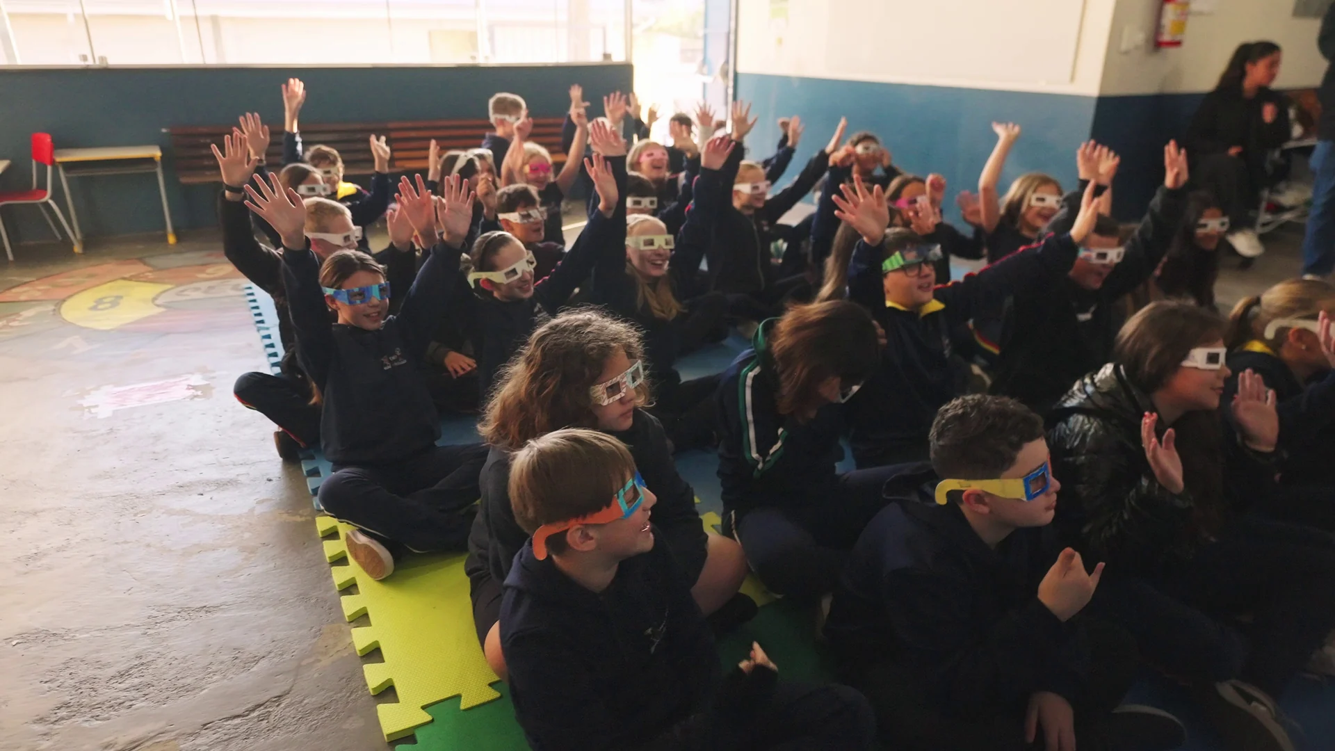 Crianças de uniforme escuro e óculos 3D levantam as mãos, sentadas no chão de uma sala de escola.