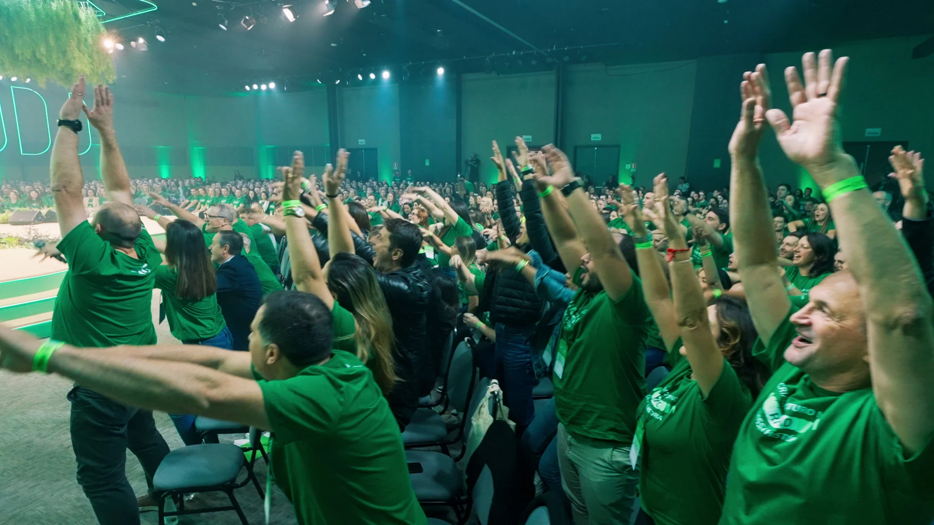 Colaboradores de camiseta verde levantam os braços e vibram num grande auditório iluminado em verde, durante o Workshop de 40 anos da Sicredi Serrana.