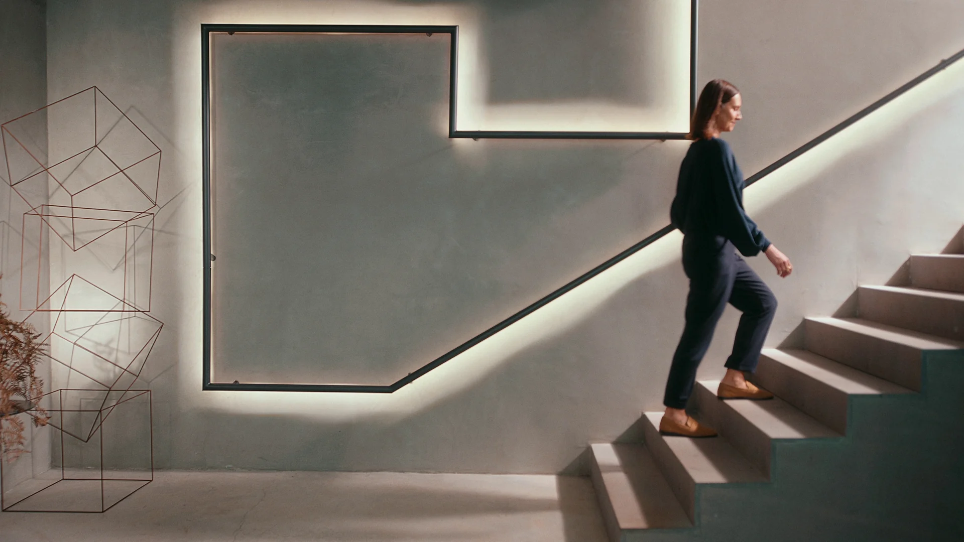 Mulher de roupa escura sobe uma escada de concreto contornada por uma linha de luz na parede, numa casa de arquitetura contemporânea.