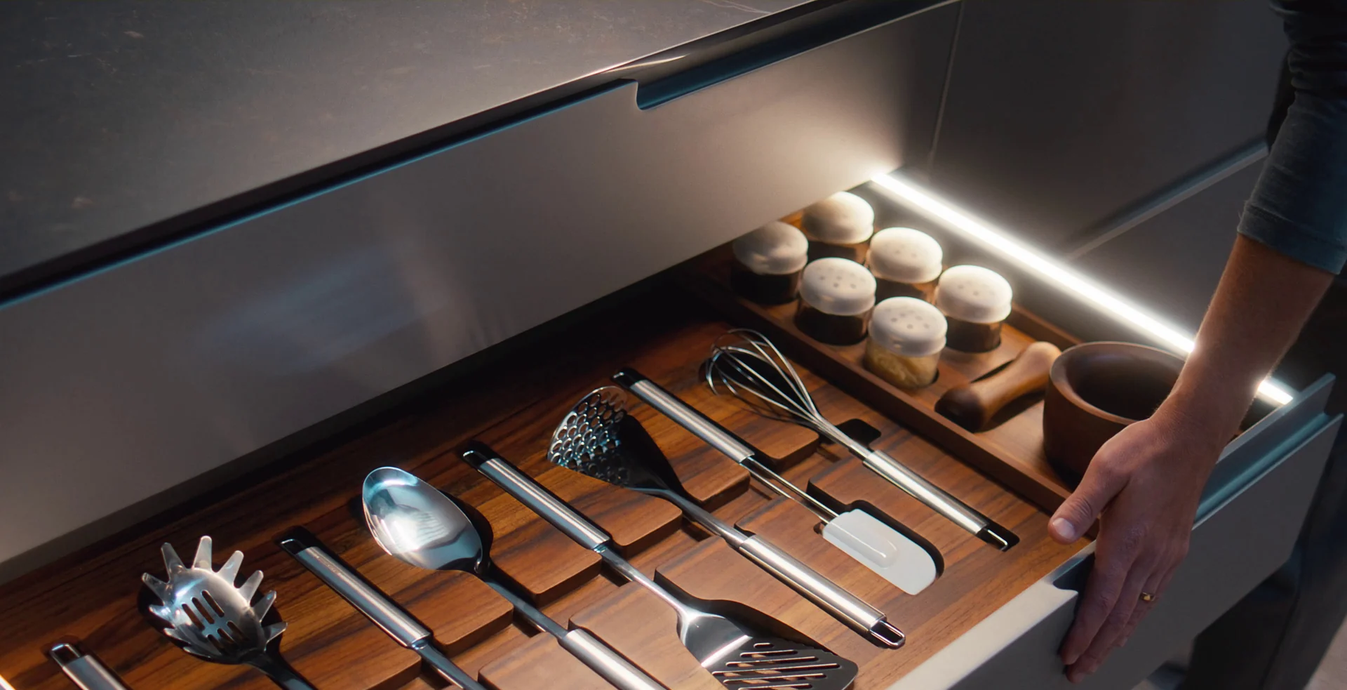 Mão fecha uma gaveta de cozinha com fita de LED, que organiza espátulas, concha, batedor e potes de tempero em divisórias de madeira escura.