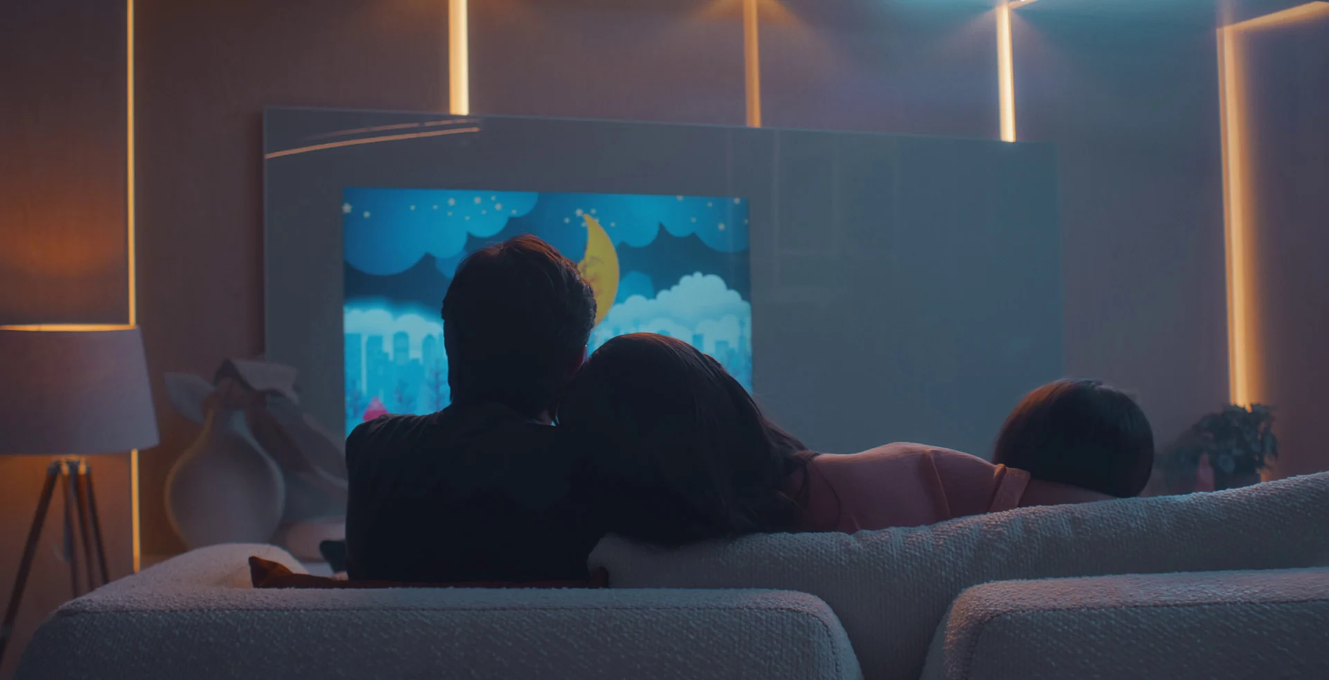 Pai, mãe e filha de costas no sofá assistem a um desenho animado de lua e nuvens numa TV embutida em painel com iluminação de LED, em sala à noite.