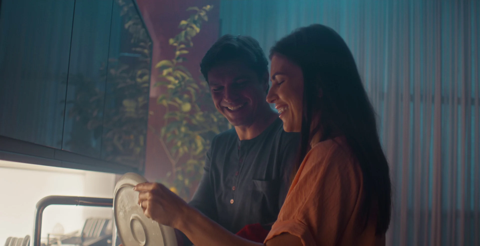 Pai e mãe sorriem enquanto lavam e secam uma tampa na pia da cozinha, numa luz azulada de fim de noite.
