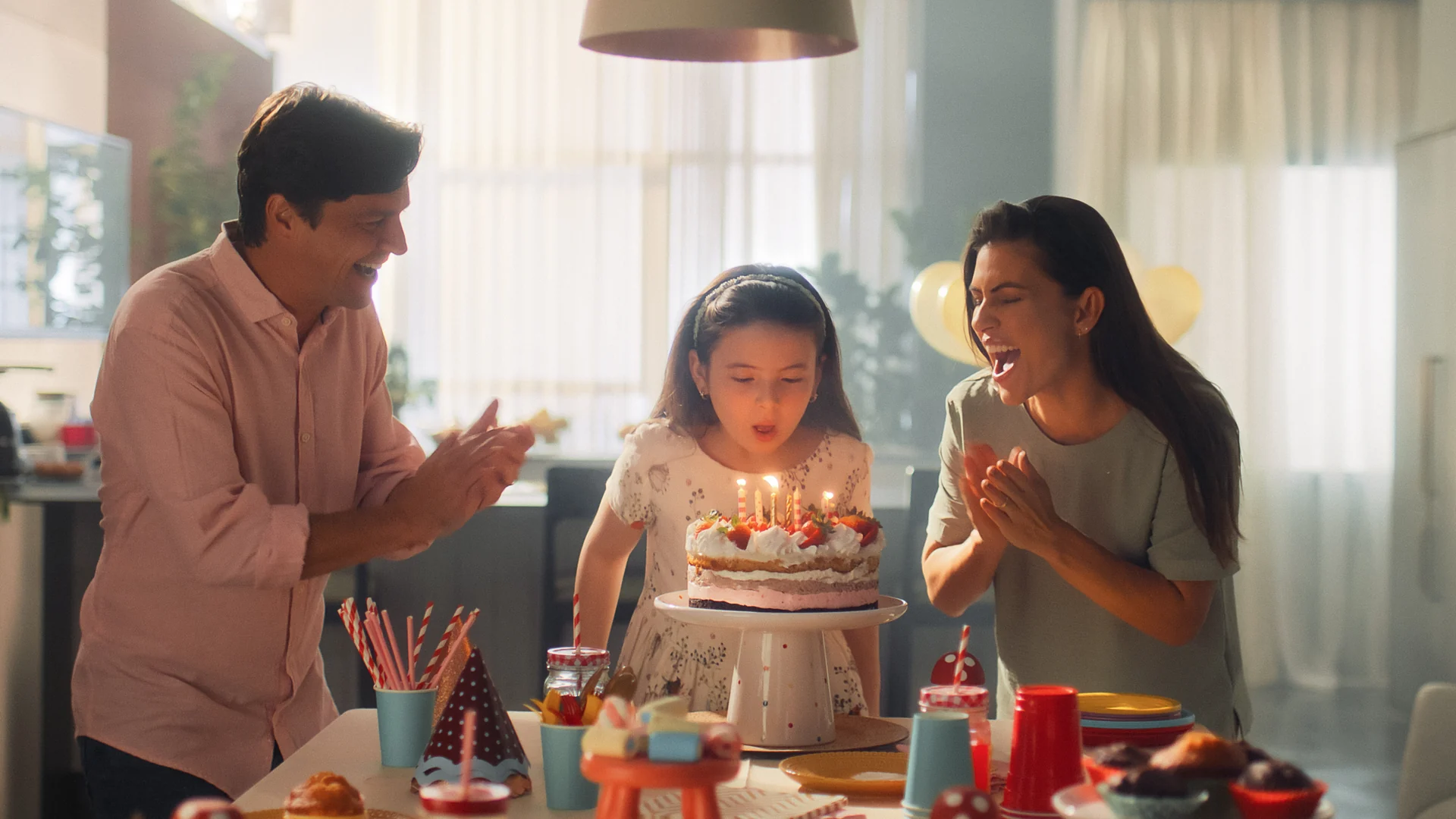 Pai, menina e mãe numa cozinha clara reunidos em torno de um bolo com velas acesas; a menina sopra as velas enquanto os pais sorriem e aplaudem.