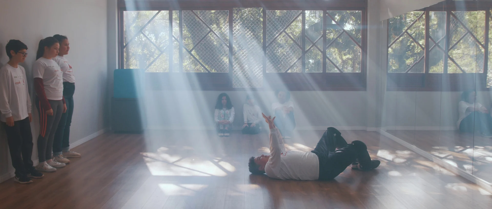 Feixes de sol entram pelas janelas de uma sala de dança; um aluno deitado no chão ergue o braço enquanto colegas observam encostados na parede.