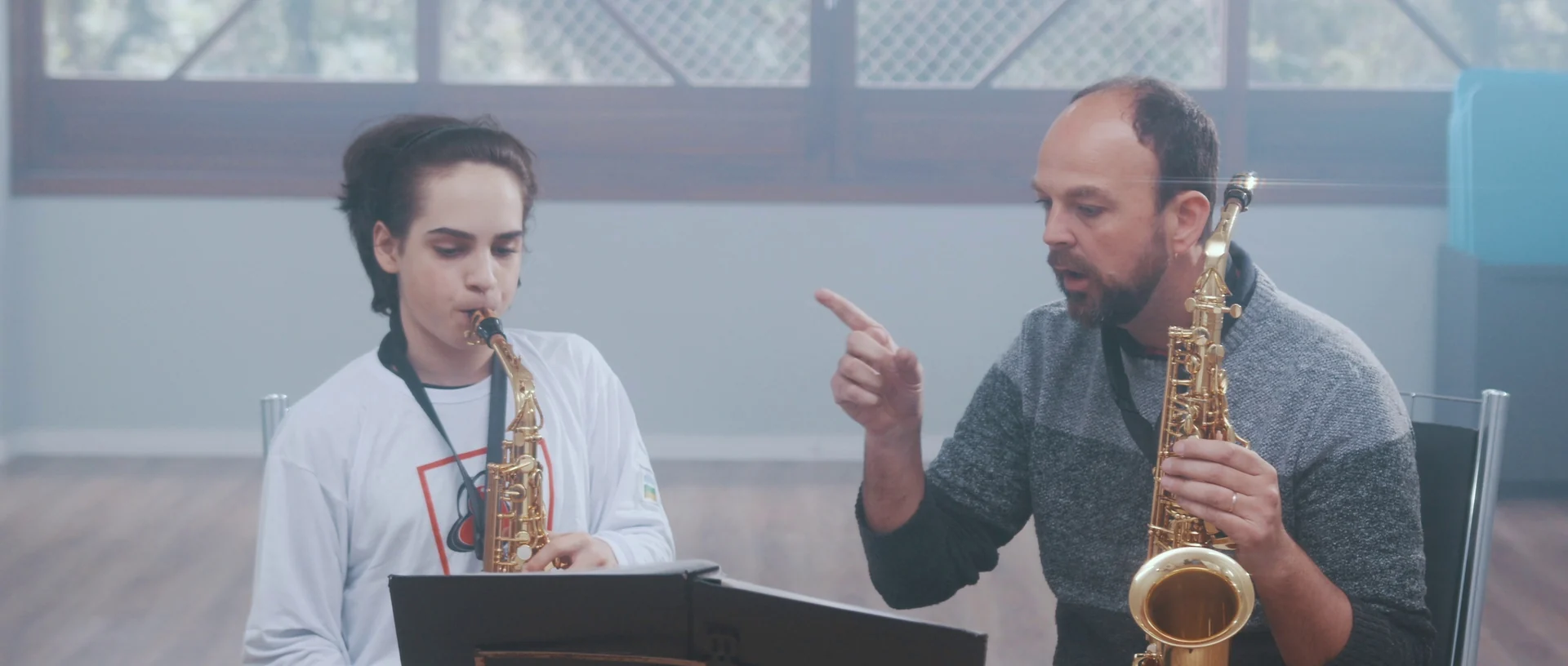 Professor com saxofone aponta e orienta um aluno que toca saxofone diante de uma estante de partitura.