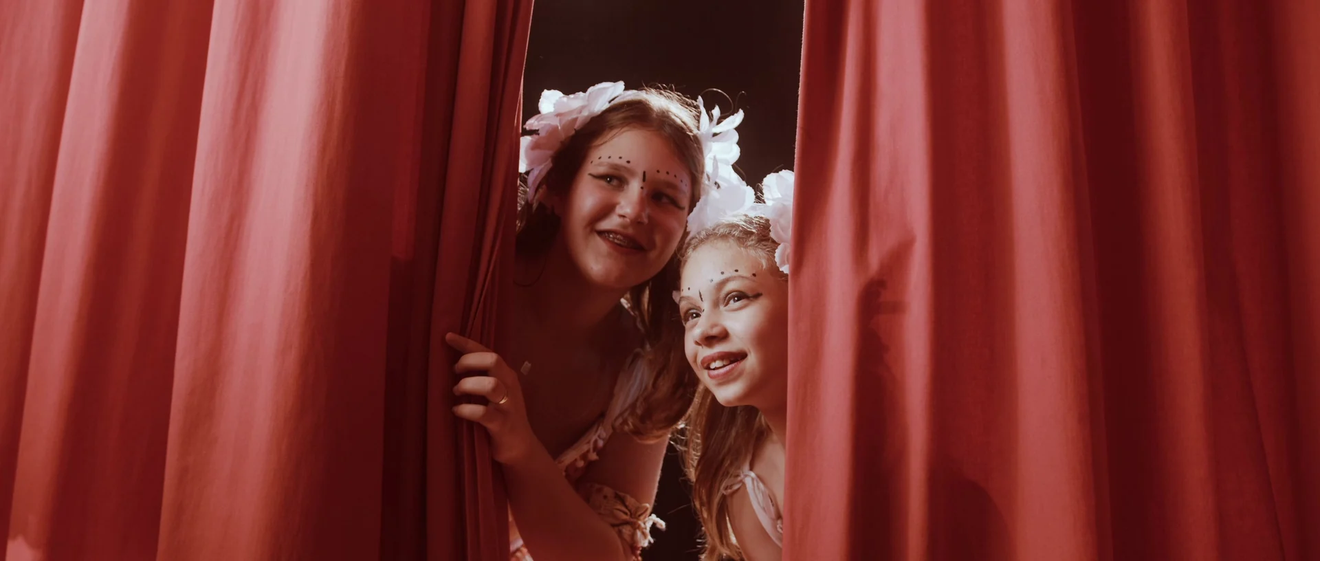 Duas meninas com flores no cabelo e maquiagem de espetáculo espiam sorrindo por uma fresta da cortina vermelha.