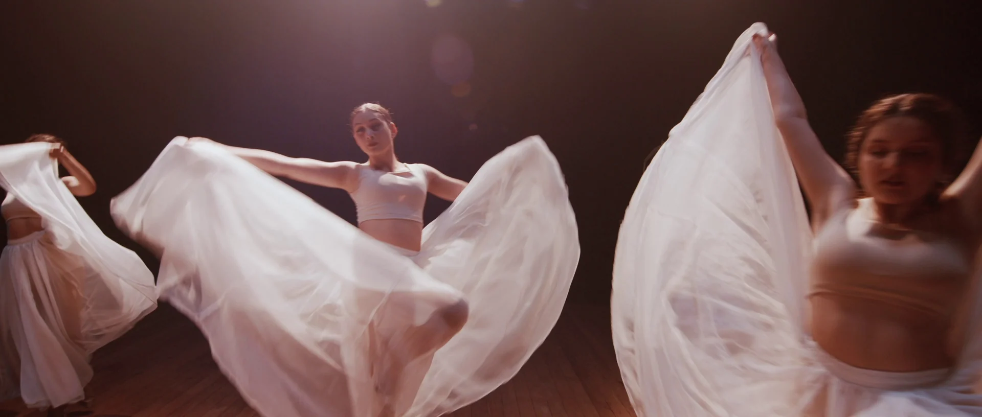 Bailarinas giram longas saias brancas sobre o palco escuro, em movimento.
