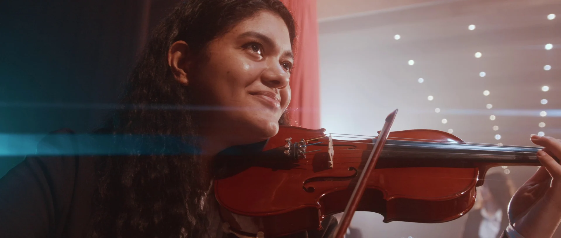 Jovem violinista sorri enquanto toca, com as luzes do teatro desfocadas ao fundo.