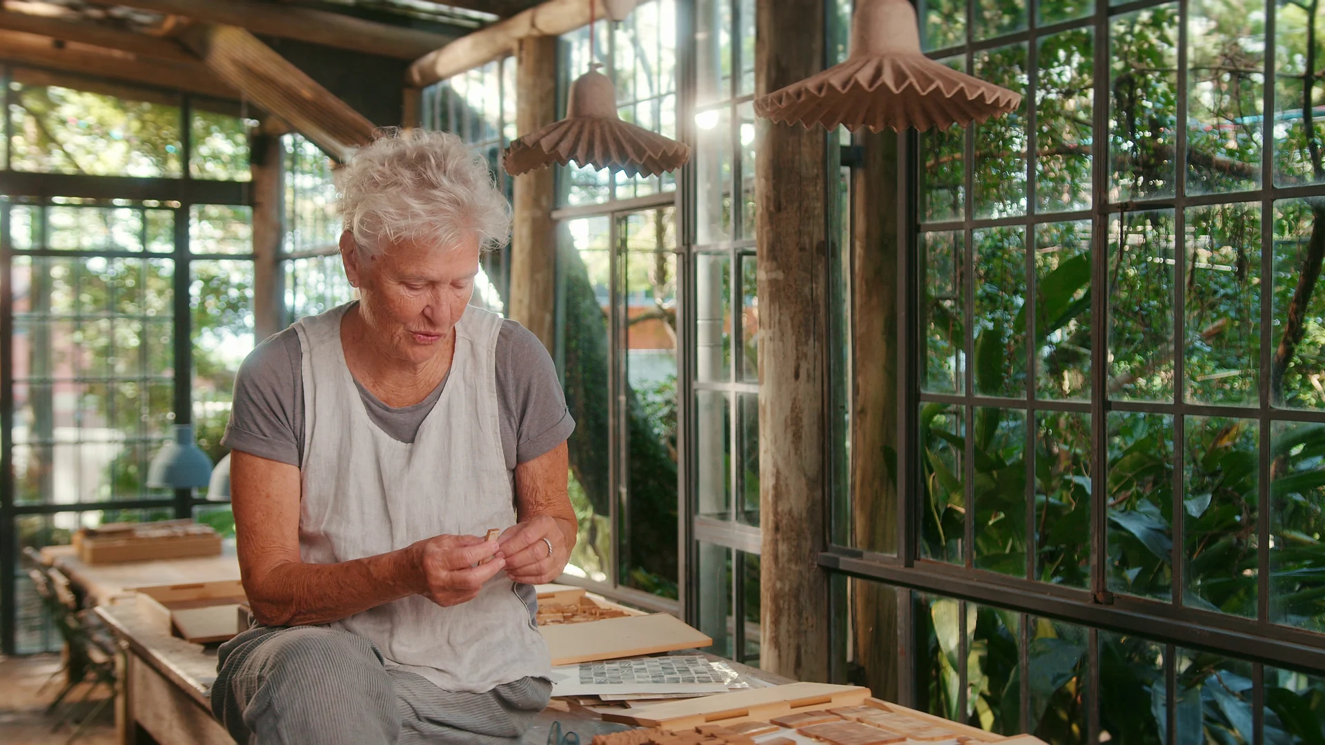 Heloisa Crocco, de avental, manuseia uma pequena peça de madeira sentada à mesa de trabalho de um ateliê envidraçado, cercado de vegetação e luminárias pendentes.