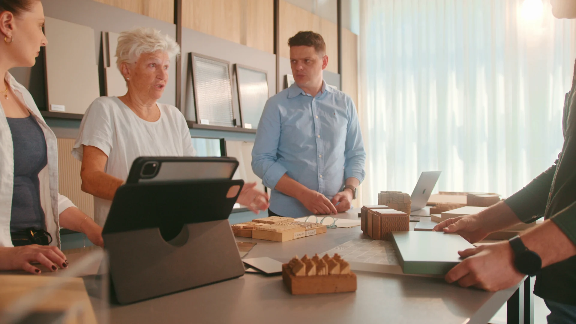 Heloisa Crocco conversa com integrantes das equipes em torno de uma mesa com tablet e amostras de madeira, em um showroom claro.