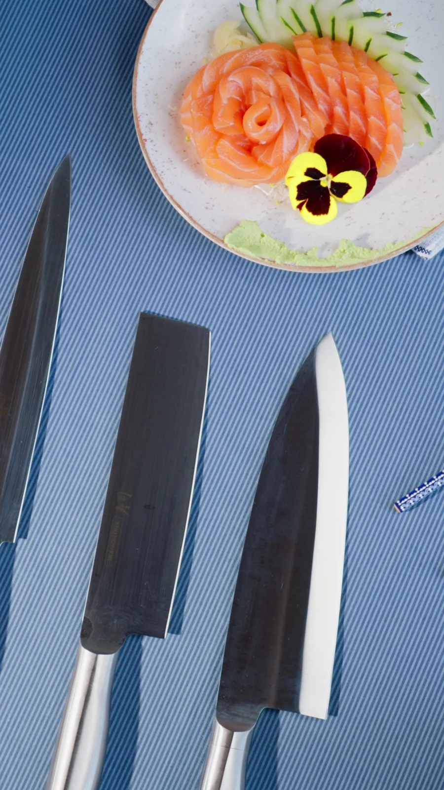 Facas da linha Sushi alinhadas sobre fundo azul, com prato de sashimi de salmão e flor comestível.