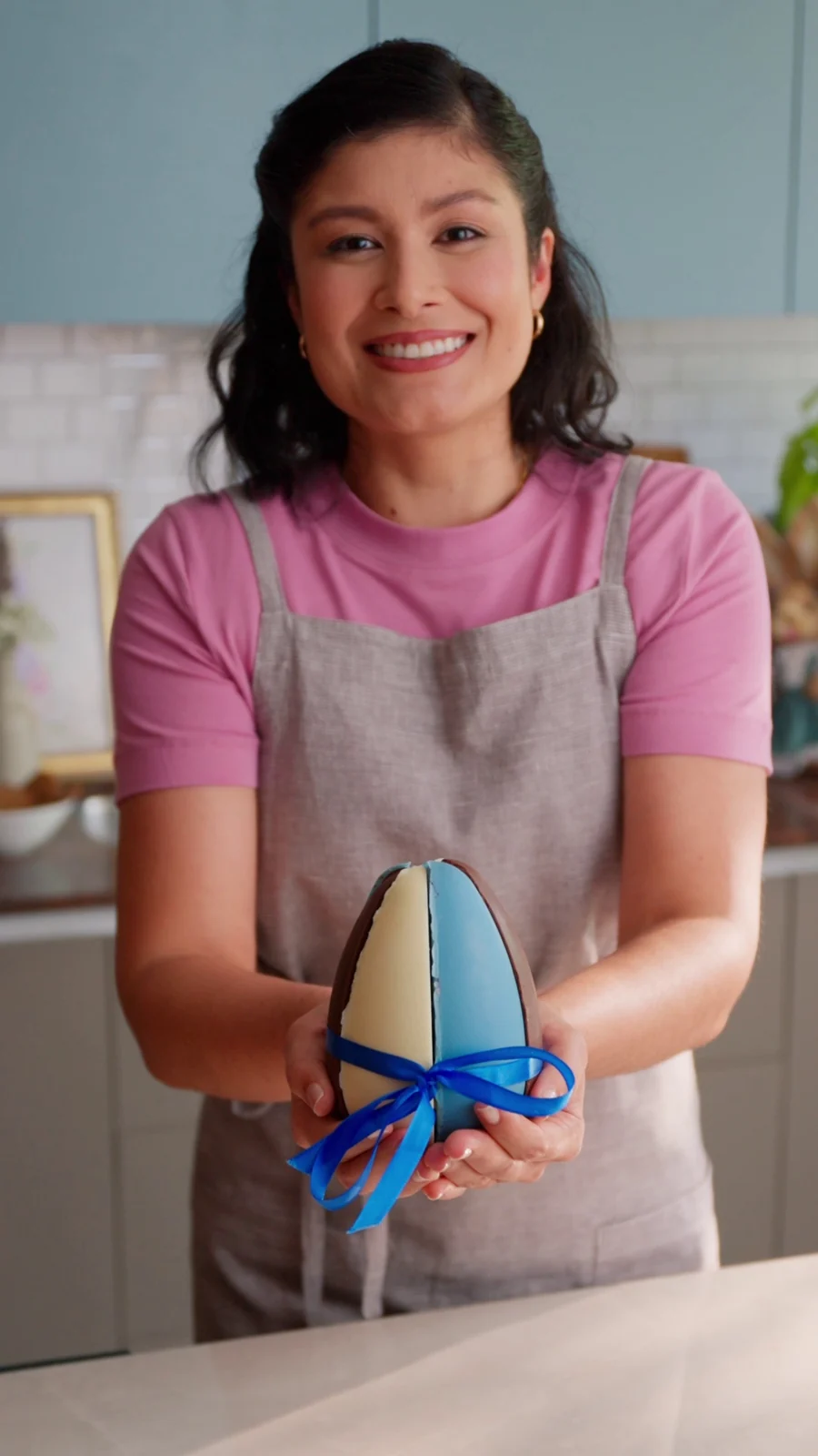 Culinarista de avental sorri e apresenta um ovo de Páscoa de fatia amarrado com fita azul.