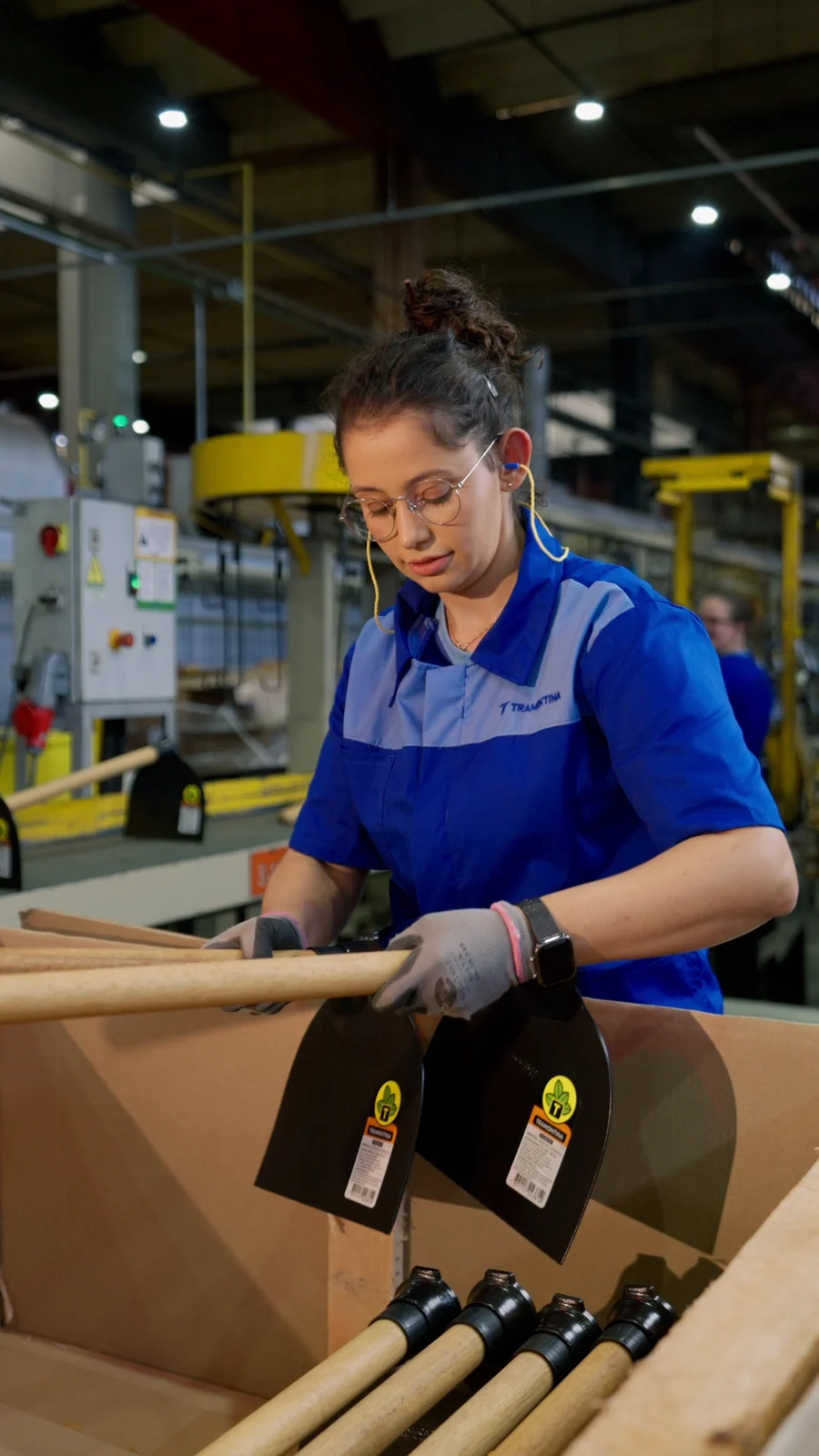 Colaboradora de uniforme azul embala enxadas Tramontina na linha de produção da fábrica.