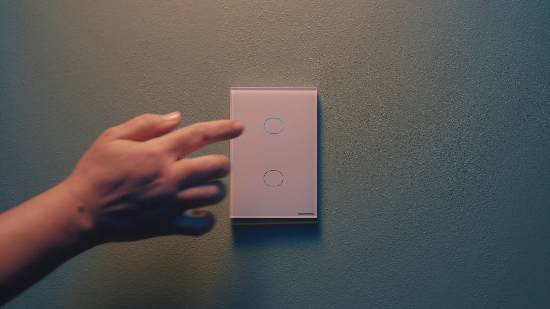 Mão se aproxima de um interruptor touch rosado de duas teclas circulares em uma parede de tom azulado.