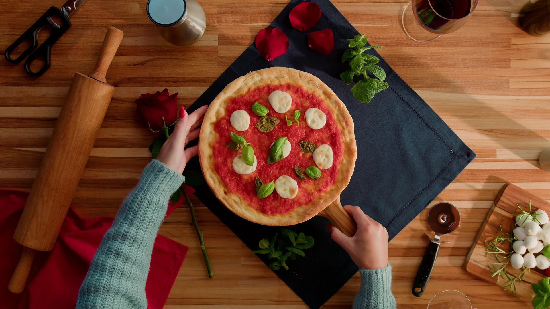 Pizza margherita vista de cima sobre pá de madeira, com rosa vermelha, rolo de massa e utensílios na bancada.