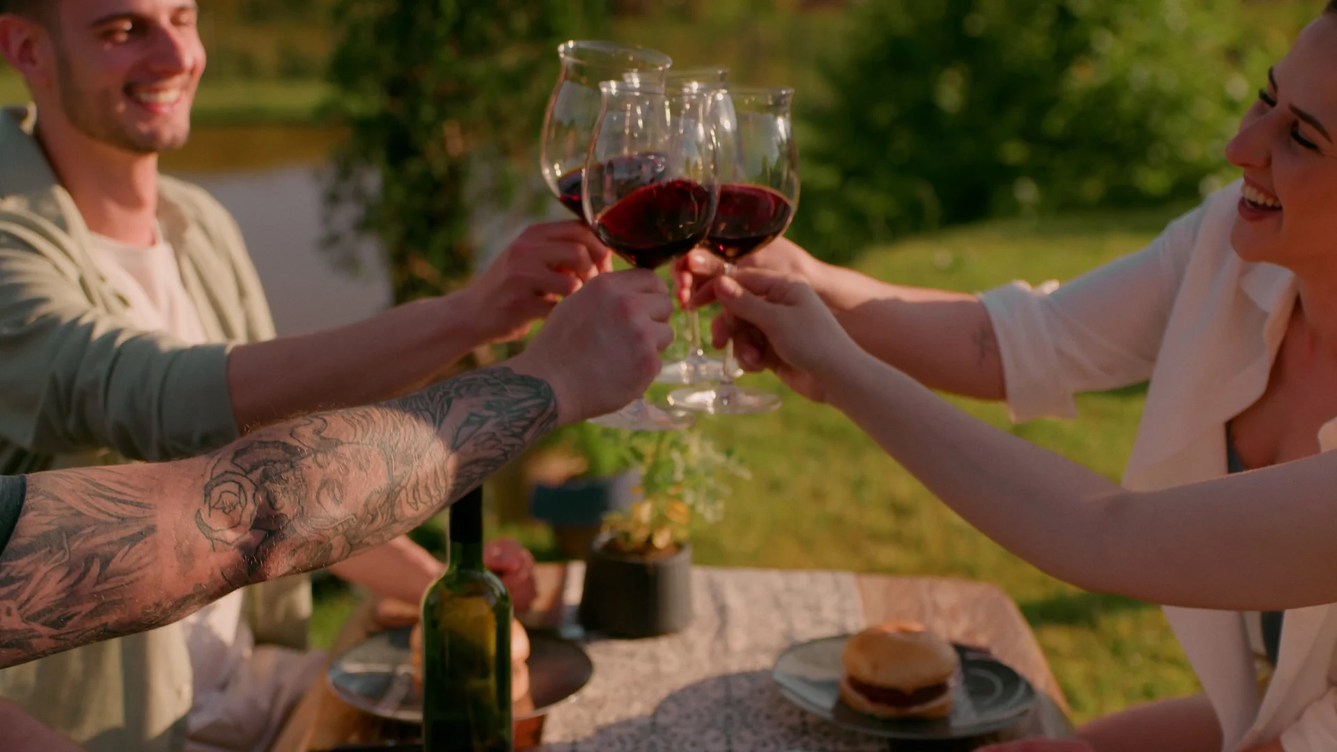 Amigos sorridentes brindam com taças de vinho tinto em uma mesa ao ar livre, sob sol de fim de tarde.