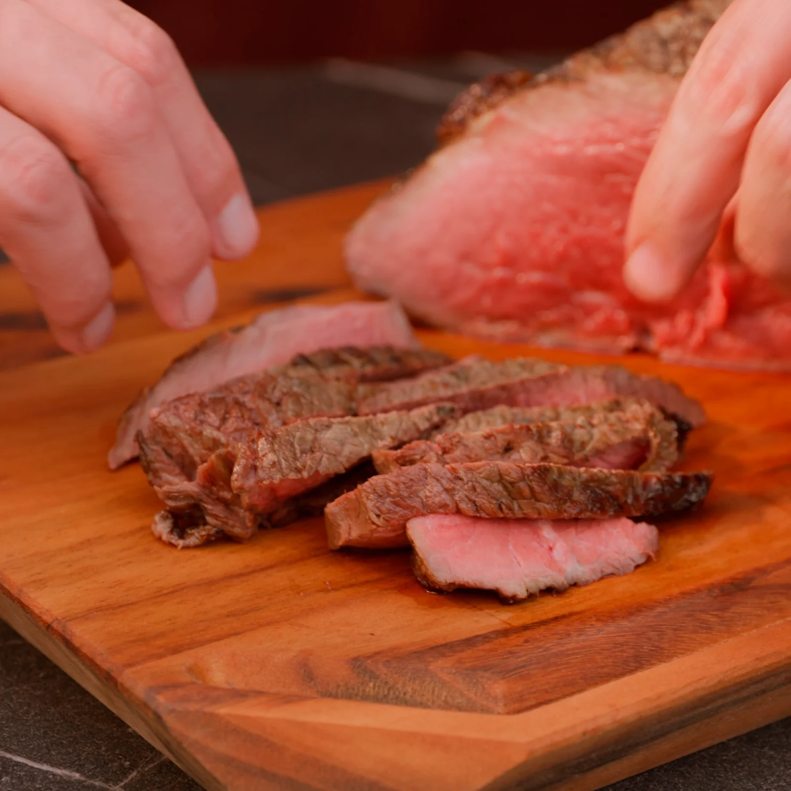 Fatias de carne assada, rosadas por dentro, sobre a tábua de madeira, com as mãos do cozinheiro ao fundo.