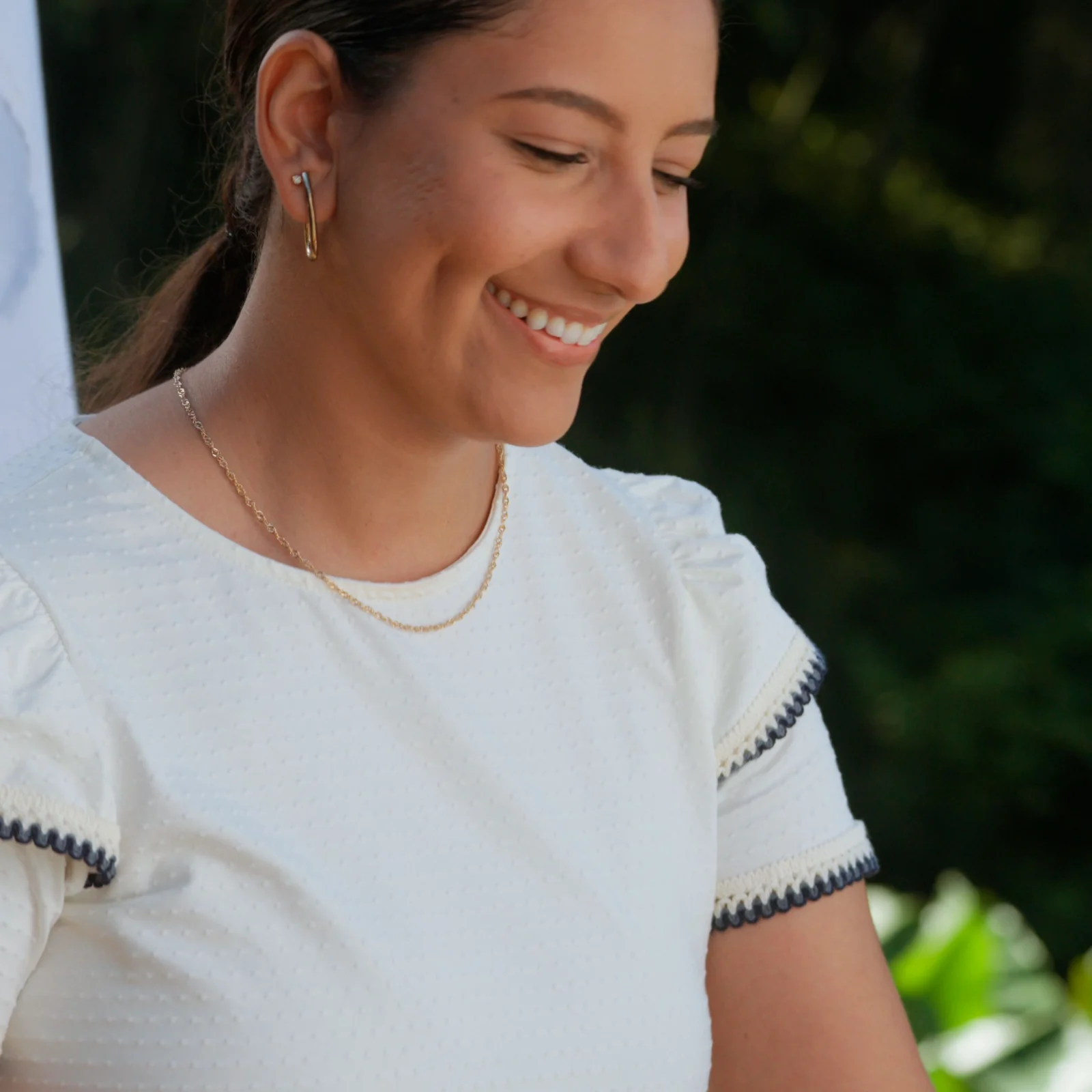 Mulher de blusa branca com detalhes azuis sorri olhando para baixo, com folhagens desfocadas ao fundo.