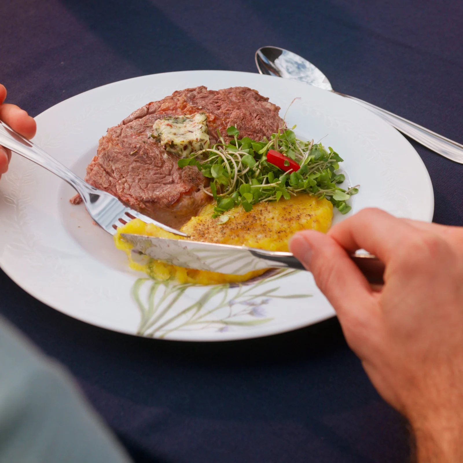 Garfo e faca do faqueiro Laguna cortam um bife com manteiga de ervas e salada de brotos em prato branco.
