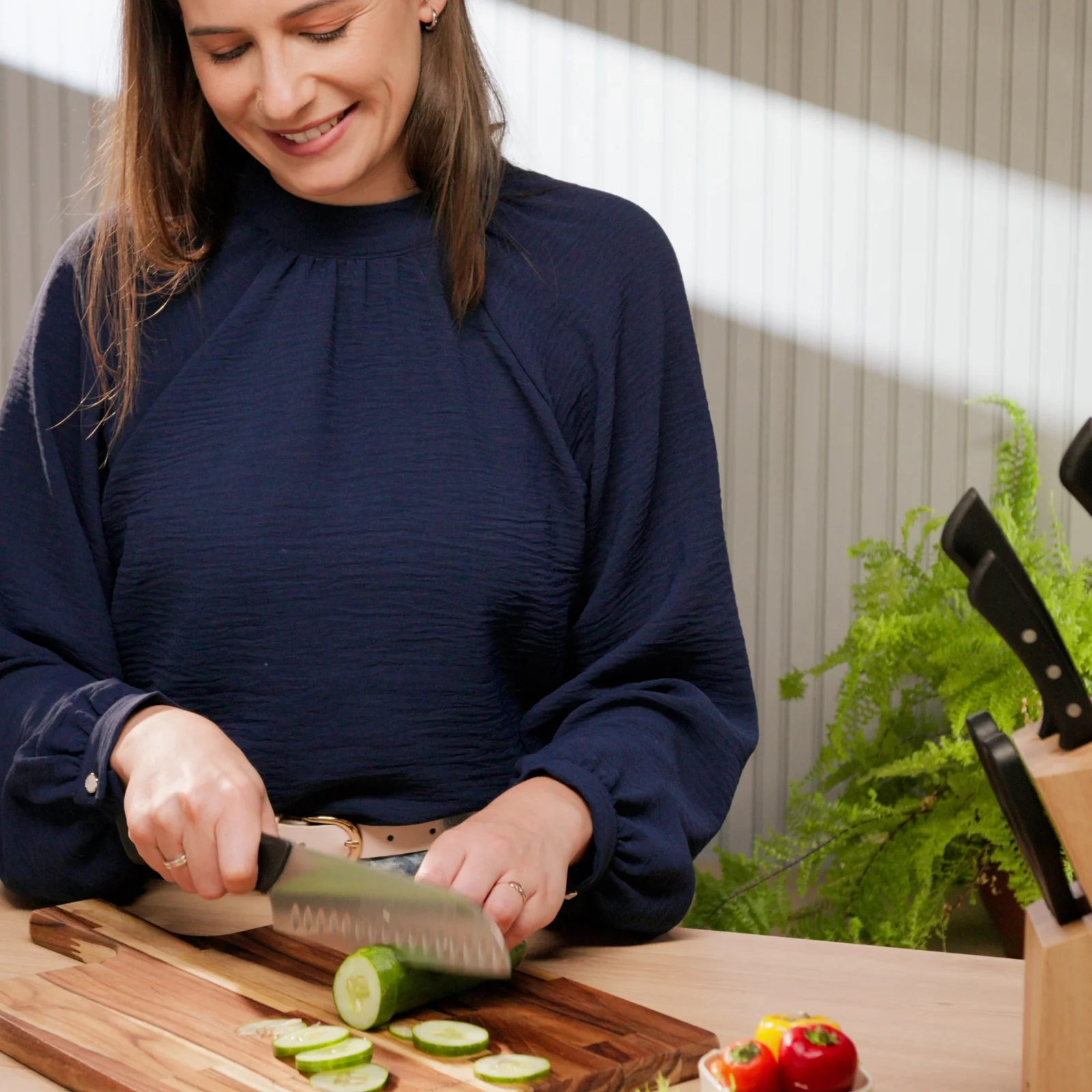 Mulher sorridente corta pepino com a faca Santoku sobre tábua de madeira, com bloco de facas ao lado.