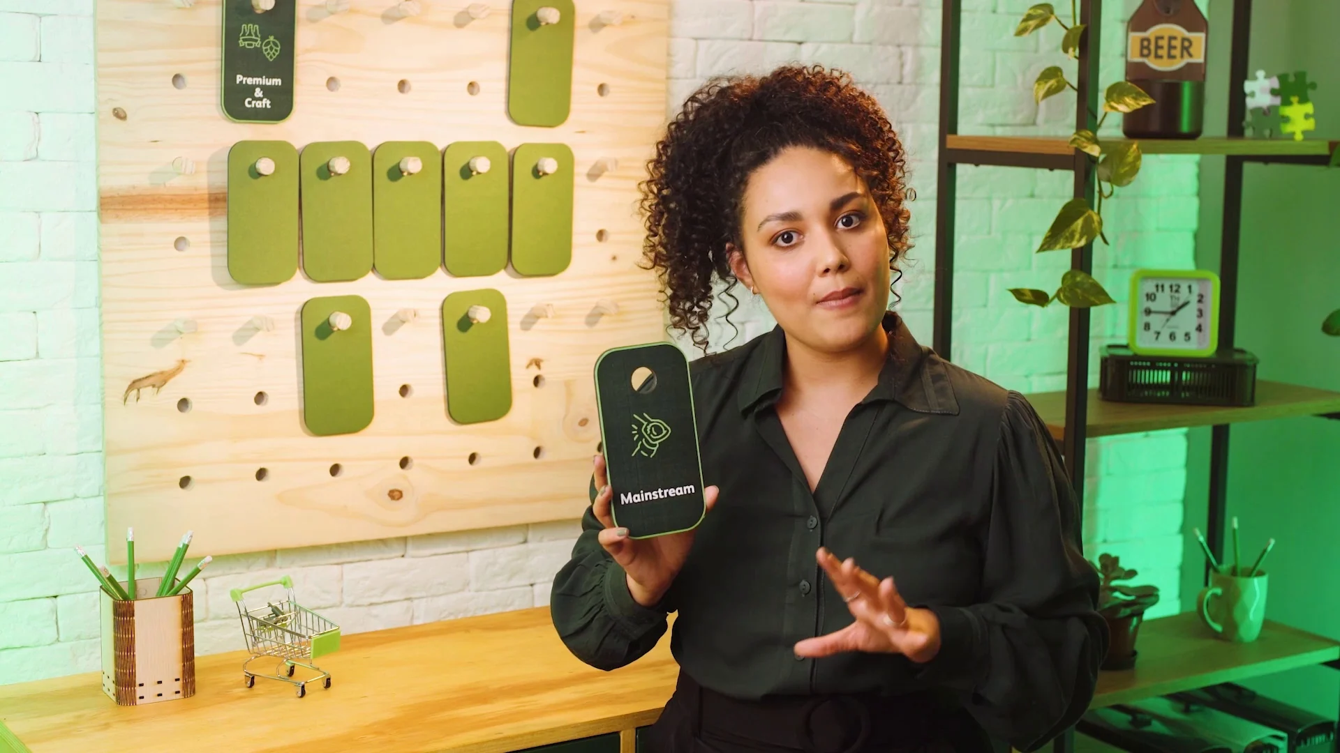 Apresentadora diante de um painel de pegboard com cartões verdes segura um cartão de categoria de marca e gesticula.