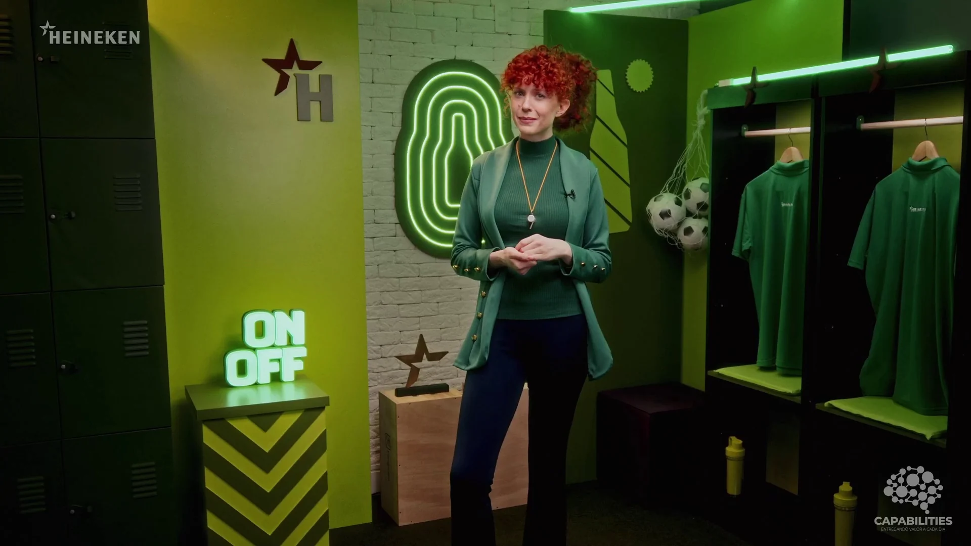 Apresentadora de pé em um vestiário cenográfico com camisas verdes penduradas, bolas de futebol e um totem luminoso com as palavras ON e OFF.