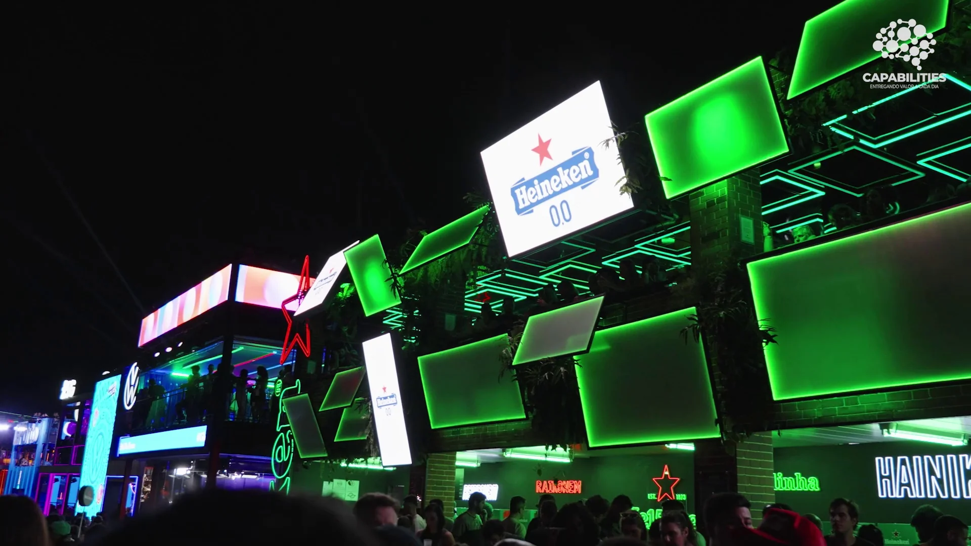 À noite, no festival The Town, a fachada do bar da Heineken brilha com painéis e molduras de neon verde sobre a multidão, com um telão da Heineken 0.0 no alto.