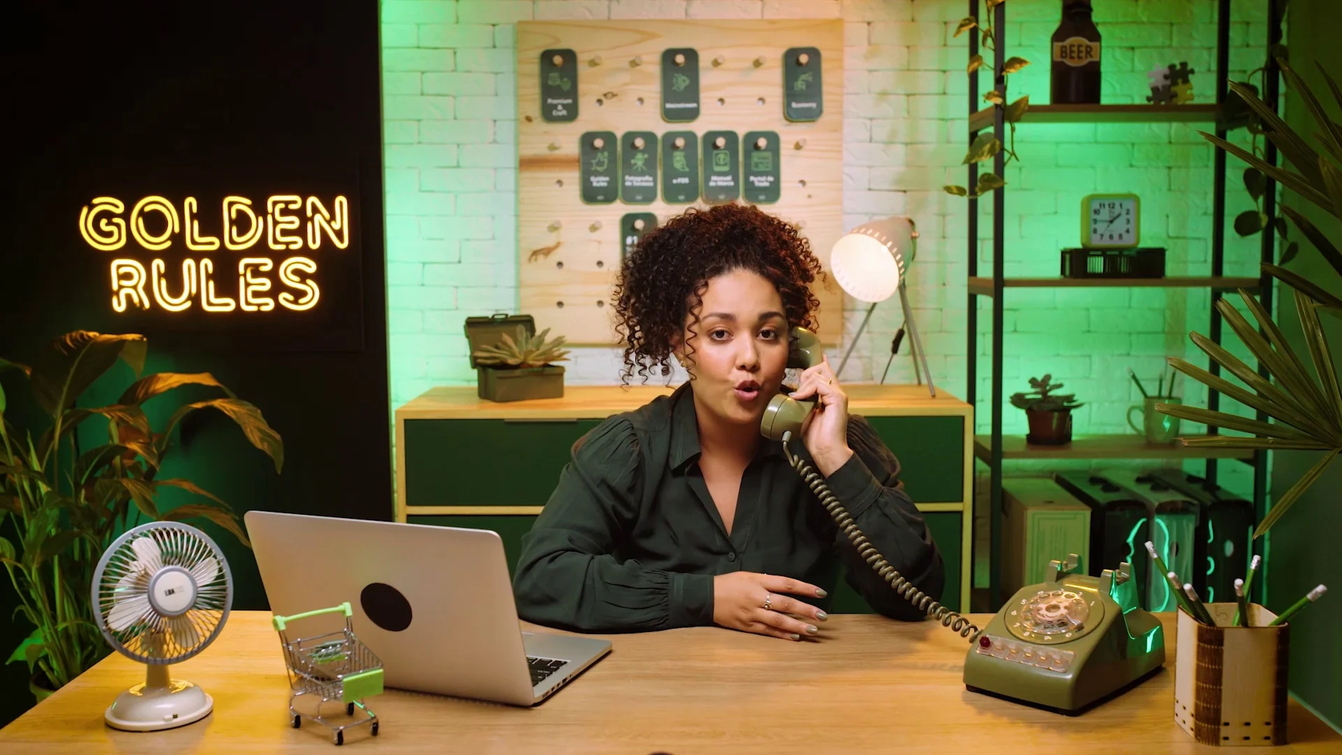 Apresentadora de cabelo cacheado e blusa verde-escura atende um telefone de disco verde numa mesa de madeira, com notebook, ventilador retrô e o neon Golden Rules ao fundo, sobre parede de tijolos iluminada em verde.